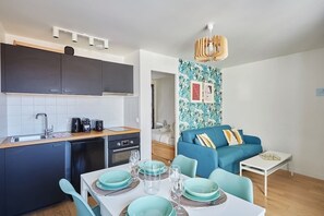 Una cocina moderna con armarios oscuros, lavavajillas y cafetera. Hay un comedor con una mesa preparada para dos personas, con platos y vasos turquesa. Se ve un sofá azul con almohadones decorativos en la sala de estar.