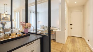 A modern kitchen with a black countertop, white cabinets, and a large mirror. There is a dining area with a white table and chairs, and a small round table with a vase of flowers. The room has wooden flooring and white walls. - Superb apartment Invalides Eiffel Tower (Paris)