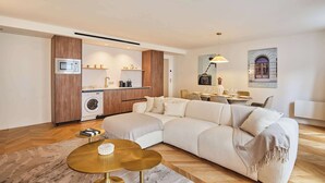 Ein modernes Wohnzimmer mit einem Ecksofa, einem kleinen runden Tisch, einer Waschmaschine und einer Küchenzeile mit Mikrowelle und Regalen.