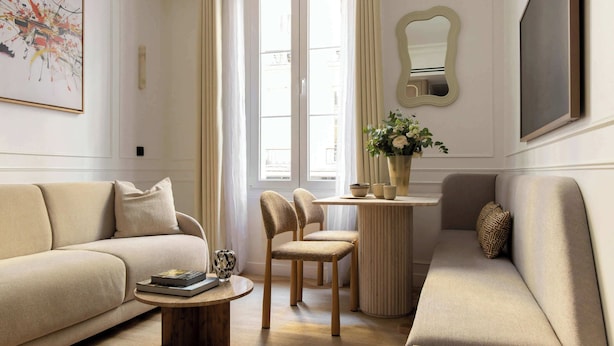 Un salon avec un canapé beige, une table basse et deux fauteuils. Il y a une fenêtre avec des rideaux, un vase avec des fleurs et un tableau encadré au mur.