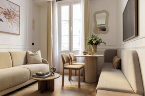 A living room with a beige sofa, a coffee table, and two chairs. There is a window with curtains, a vase with flowers, and a framed picture on the wall. - Stunning apartment with AC - Center of Paris (Paris)