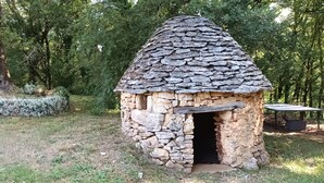 Une construction en pierre, surmontée d’un toit de chaume, et entourée de verdure.