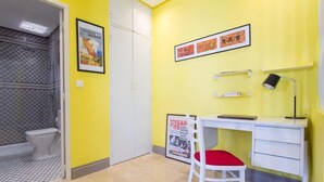 Ein Zimmer mit gelber Wand, einem weißen Schreibtisch mit Stuhl, einem gerahmten Plakat, einer schwarzen Lampe und einem gefliesten Duschbereich.