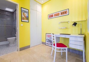 Ein Zimmer mit gelber Wand, einem weißen Schreibtisch mit Stuhl, einem gerahmten Plakat, einer schwarzen Lampe und einem gefliesten Duschbereich.