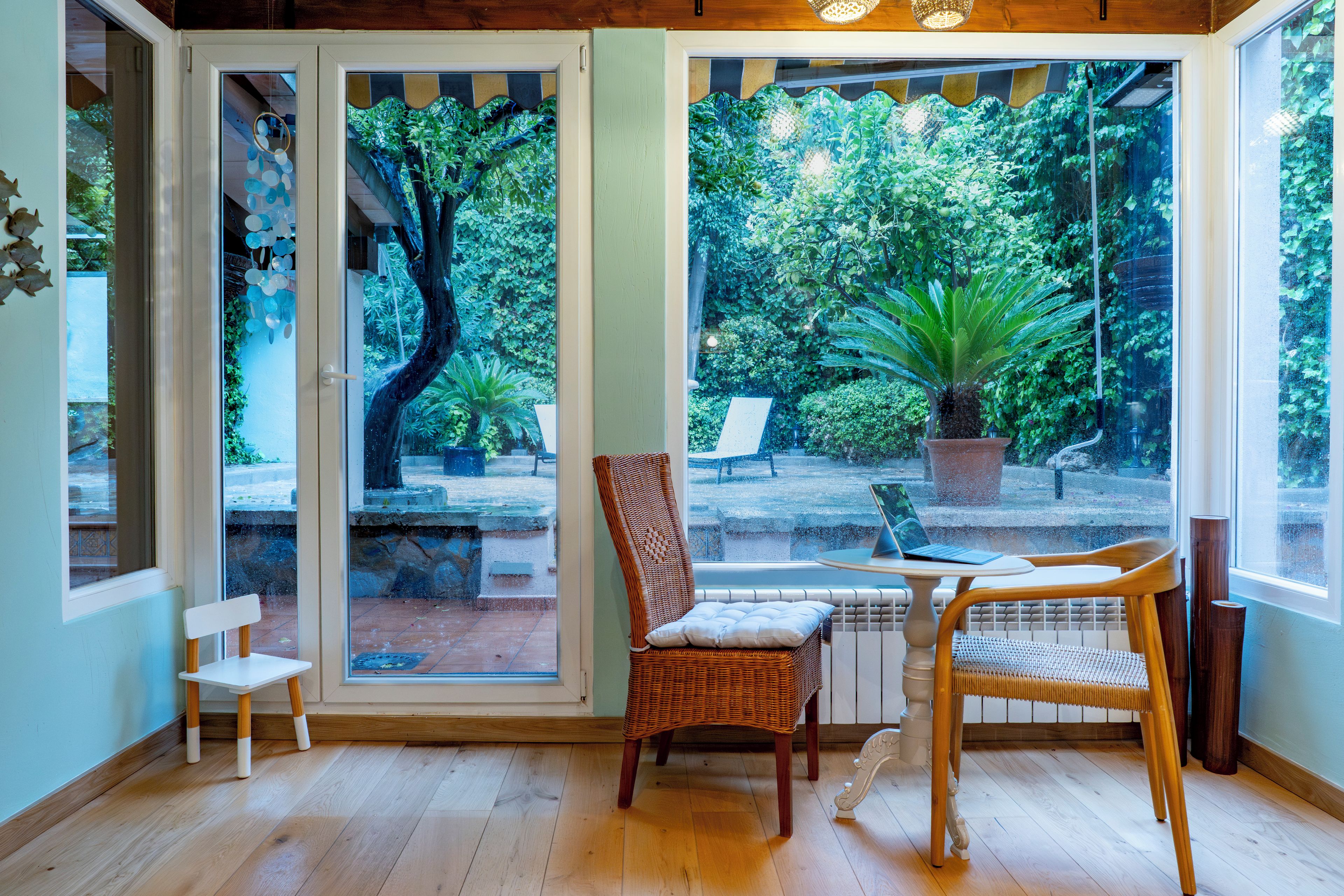 A room with a view of a garden through glass doors, a wicker chair, a small table, and a white chair.
