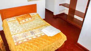 Un lit avec une couvre-lit à motifs, un oreiller et un mouchoir blanc, le tout placé devant une tête de lit en bois, dans une pièce dont le sol est rouge et les murs blancs.
