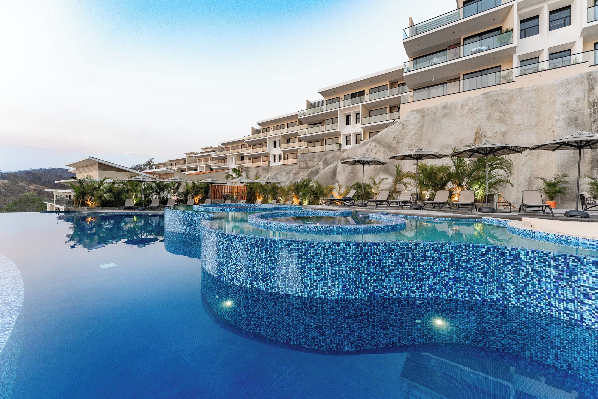 A modern hotel with a large infinity pool, outdoor seating, and a view of the surrounding landscape.