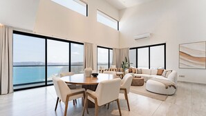 Ein geräumiges Wohnzimmer mit einem großen runden Tisch, bequemen Sitzgelegenheiten und Meerblick durch raumhohe Fenster.