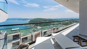 Ein Balkon mit Glasgeländer mit Blick auf eine Küstenlandschaft mit grünen Hügeln und einem Gewässer.