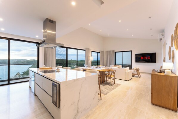 Ein geräumiger Wohnbereich mit einer großen Kücheninsel, einem Esstisch und einer Sitzecke inklusive Sofa und Sesseln. Ein Flachbildfernseher ist an der Wand befestigt, und ein großes Fenster bietet Blick auf das Meer.