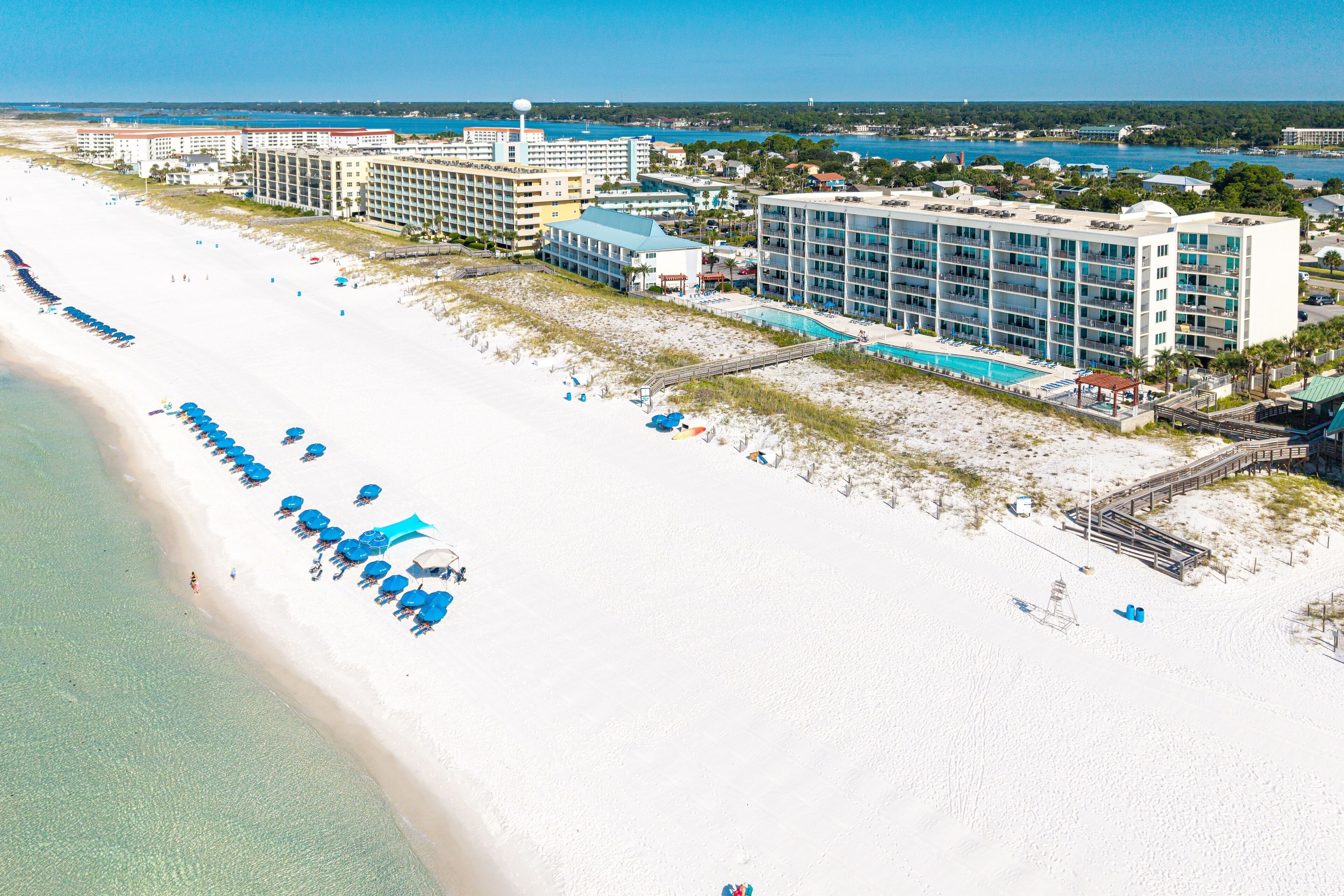 Vue aérienne d’un complexe hôtelier en bord de mer, avec une longue plage de sable blanc, une rangée de parasols bleus et un grand bâtiment d’hôtel doté de nombreux étages et de balcons.