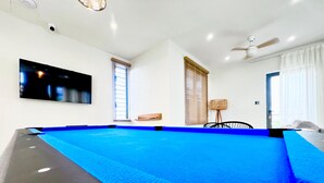 Una mesa de billar en una habitación con paredes blancas, un ventilador de techo y un televisor montado.