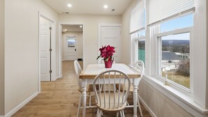 A dining area with a wooden table and white chairs, a vase with flowers, and a window with a view of buildings and trees. - Heishman Historic Hillside Home 4 Bedroom Home (Wardensville)