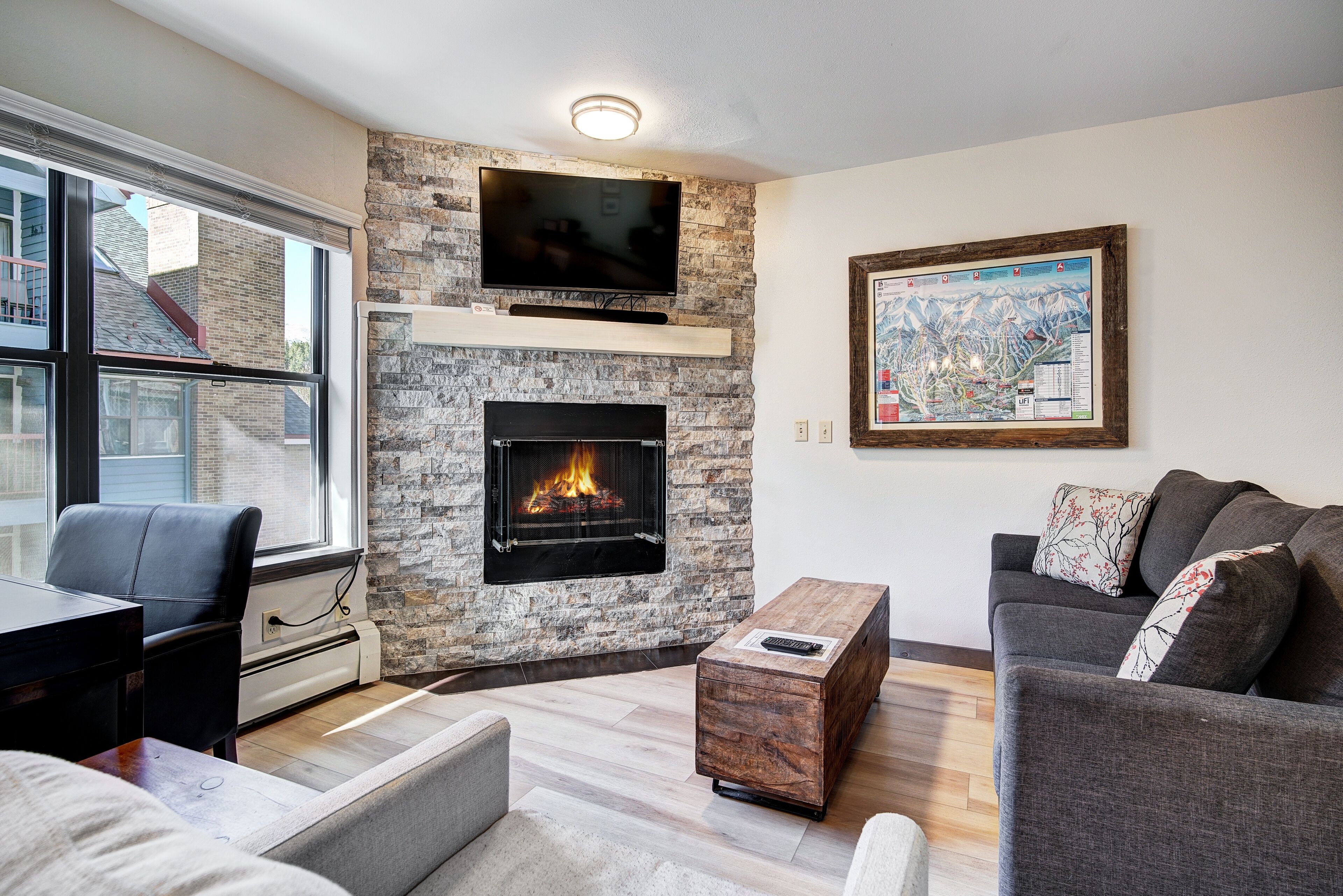 A living room with a stone fireplace, a flat-screen TV mounted above it, a map on the wall, and a sofa with cushions.
