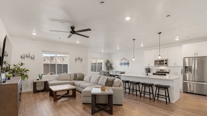 A spacious living area with a large L-shaped sofa, a small coffee table, and a kitchen area with a refrigerator, microwave, and bar stools. - Chill And Grill Getaway 4 Bedroom Townhouse (Washington)