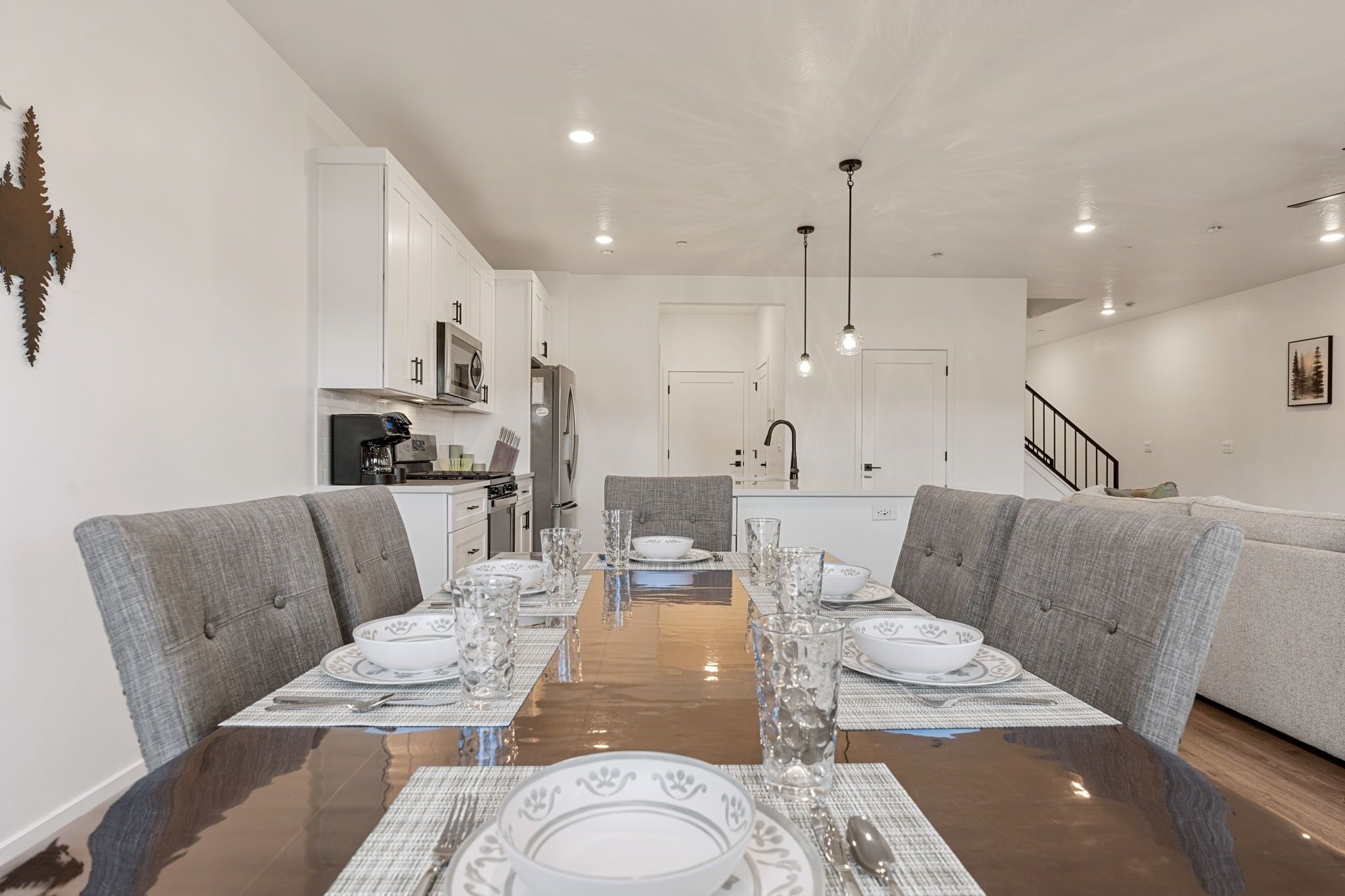 A modern dining area with a glass-top table set for six, grey upholstered chairs, and a kitchen in the background.