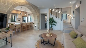 Un vaste espace de vie agrémenté d’une arche en pierre, d’une table ronde en bois, d’un canapé avec des coussins verts et d’un escalier menant à un niveau supérieur.