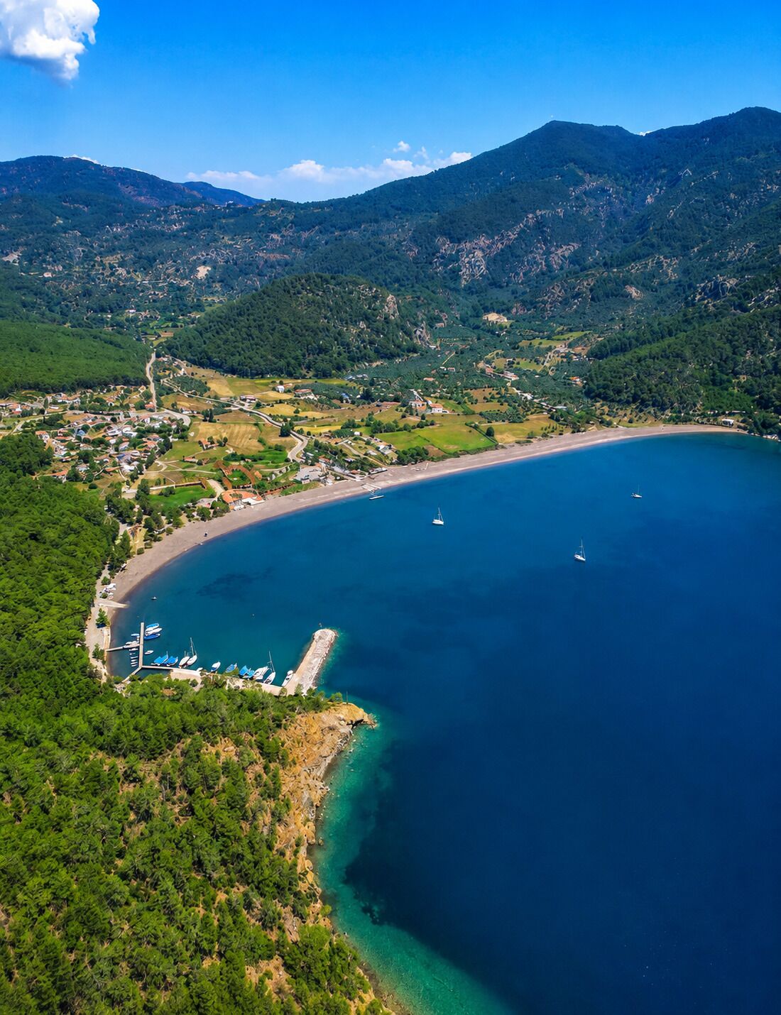 An aerial view of a coastal town with a marina, clear blue sea, and surrounding mountains.