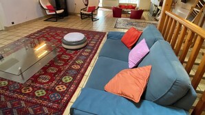 Ein geräumiges Wohnzimmer mit einem blauen Sofa, einem Teppich mit roten und blauen Mustern und einem transparenten Glastisch.