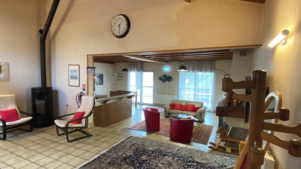 Ein geräumiges Wohnzimmer mit hoher Holzbalkendecke, einem großen Teppich und einer Couch mit roten Kissen.