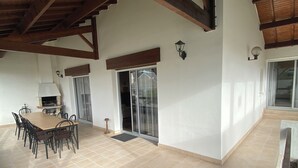 Eine geräumige Terrasse mit Fliesenboden, einem Holztisch mit Stühlen, einem eingebauten Grill und einer Glastür, die zu einem überdachten Bereich führt.