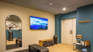 Una habitación con equipo de ejercicio, un televisor montado en la pared, una puerta y una pequeña mesa con objetos encima.