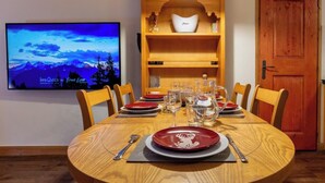 Comedor con una mesa redonda de madera dispuesta para seis personas, con platos, vasos y cubiertos. Un televisor en la pared muestra una imagen panorámica.