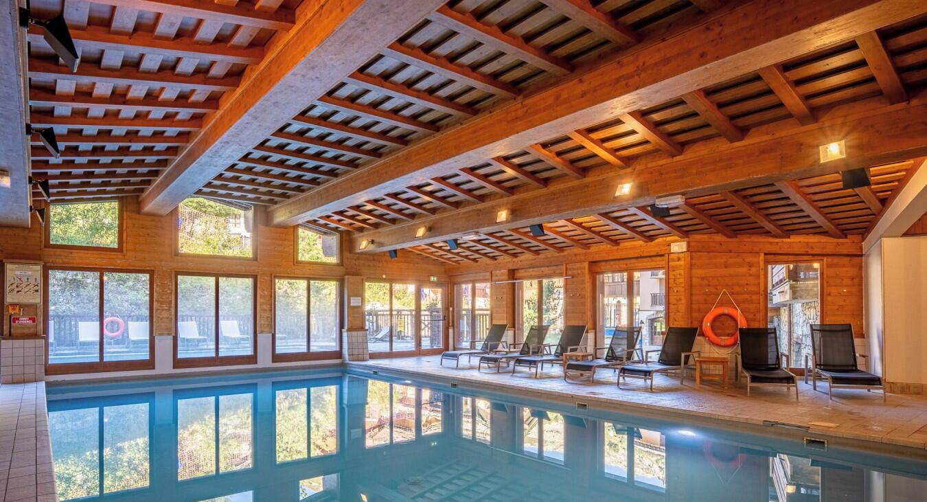 An indoor swimming pool with wooden beams, sunlit windows, and lounge chairs.