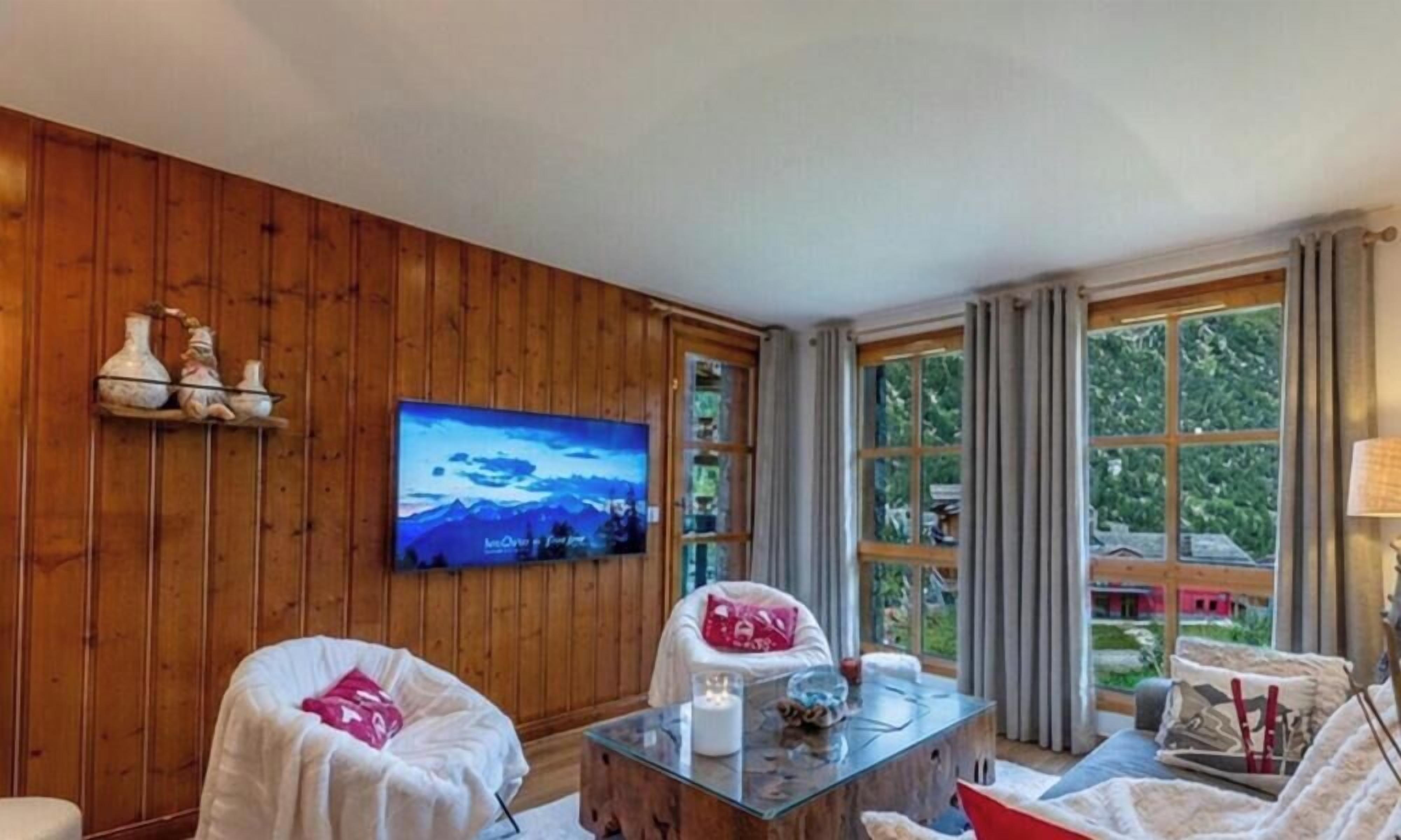 Een kamer met houten lambrisering, een aan de muur bevestigde televisie, een glazen salontafel en uitzicht op groen buiten.