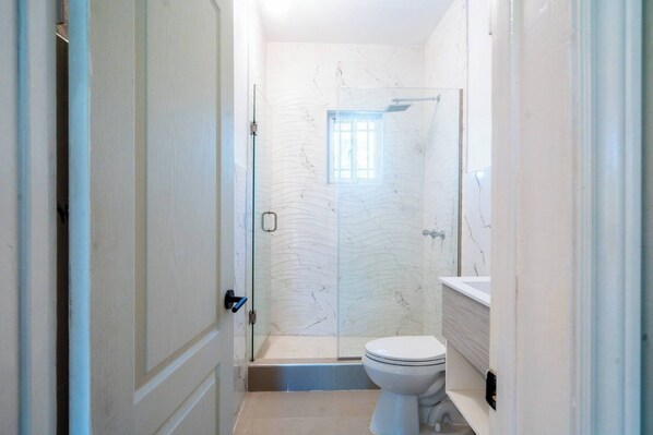 A small, narrow bathroom with a glass shower enclosure, a toilet, and a sink. - Modern Home 10minstobeach, Private Cliff, AC Wifi (Negril)