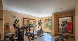 Ein Fitnessraum mit verschiedenen Trainingsgeräten, einer Wanduhr und einem Fenster mit Blick auf Grünflächen.