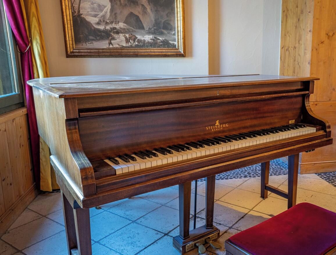 Un piano de cola con acabado de madera y el logotipo de Steinberg, ubicado en una habitación con piso de baldosas y un cuadro enmarcado en la pared.