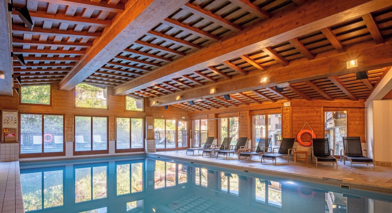 An indoor swimming pool with wooden beams, sunlit windows, and lounge chairs.