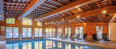 An indoor swimming pool with wooden beams, sunlit windows, and lounge chairs.