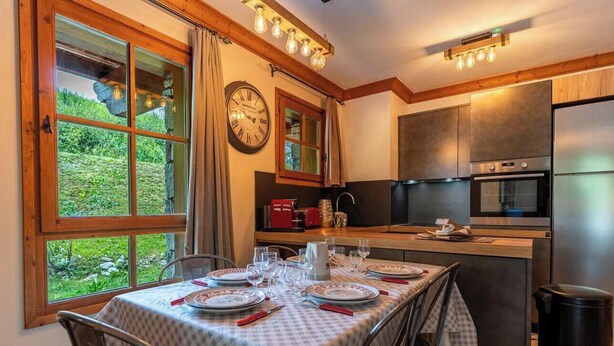 Un comedor con una mesa preparada para cuatro personas, un reloj en la pared y una cocina al fondo.