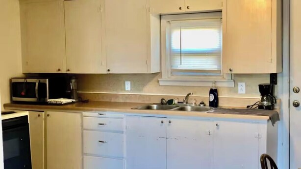 Una cocina compacta con gabinetes blancos, microondas, cafetera y ventana sobre el fregadero.