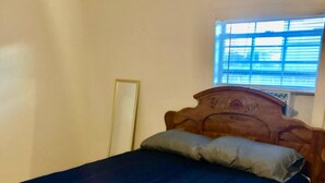 Una cama individual con colchón azul oscuro y cabecera de madera en habitación con paredes blancas y ventana con persianas.