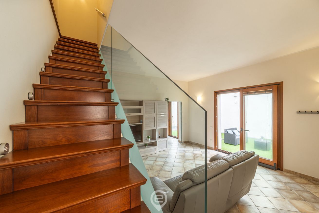 Escadaria de madeira que dá acesso a uma sala de estar com corrimão de vidro, sofá e porta de correr para área externa.