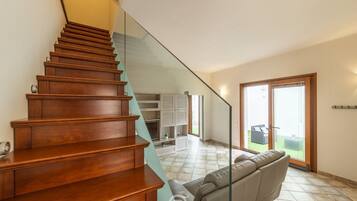 Escadaria de madeira que dá acesso a uma sala de estar com corrimão de vidro, sofá e porta de correr para área externa.
