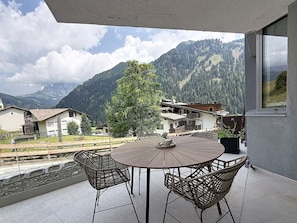 Un balcon avec une table ronde et des chaises en osier, donnant sur un paysage montagneux parsemé de maisons et traversé par une voie ferrée.