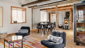 Een ruime kamer met een hoge plafond, zichtbare houten balken, een open haard, een eethoek met tafel en stoelen, en een gezellige zithoek met fauteuils en een salontafel.