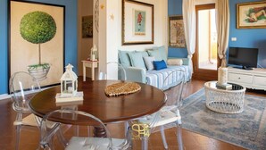 A living room with a round wooden table, clear chairs, a sofa with blue cushions, a TV, and a decorative tree on the wall. - Panoramic penthouse on the Gulf of Alghero 2 (Alghero)