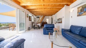 Ein modernes Wohnzimmer mit Holzbalkendecke, einem blauen Sofa, einem Glastisch und Meerblick durch eine Schiebetür.