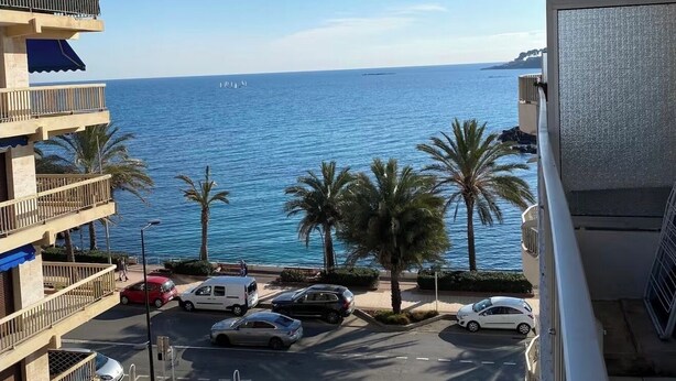 A multi-story building with balconies overlooking a street with parked cars and palm trees, leading to a body of water. - 2 rooms 8 av de l'ilette (Antibes)
