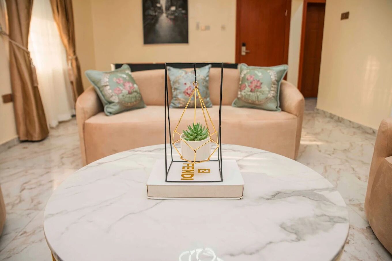 A round marble table with a gold geometric plant stand and a book. Behind, a beige sofa with floral cushions and a framed artwork on the wall.