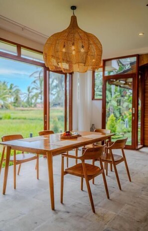 A dining area with a wooden table and chairs, a woven pendant light, and large windows offering a view of greenery. - Prana Eco Wooden Villa Ubud (Ubud)