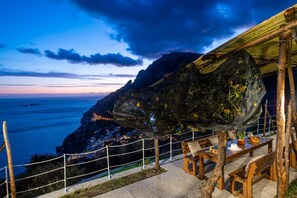 Vista costera con banco y mesa de madera bajo una zona cubierta, con vista al mar y un pueblo.