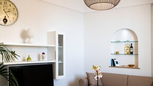 Un salon comprenant un téléviseur à écran plat, un canapé, une horloge décorative, des étagères remplies de livres et de bouteilles, et un luminaire suspendu.