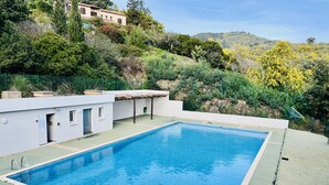 Ein rechteckiger Swimmingpool mit glasklar blauem Wasser, umgeben von einer niedrigen weißen Mauer und einem schwarzen Metallzaun. Im Hintergrund erstreckt sich eine Hügelsicht mit üppiger Vegetation und ein Gebäude mit einem Ziegeldach.
