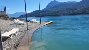 Una playa de guijarros con sillones blancos y un paseo de concreto que conduce a un lago, rodeado de montañas.